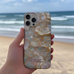 Può includere: Una custodia per telefono perlata con un design a mosaico nei toni del bianco, dell'oro e dell'iridescente. La custodia presenta un ritaglio per la fotocamera e il nome "Bella" in corsivo. Lo sfondo mostra una spiaggia e il mare.