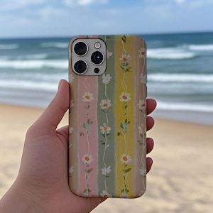 Puede incluir: Una funda de teléfono floral con un diseño de margaritas. La funda presenta rayas verticales en beige, rosa, verde y amarillo. El diseño de la margarita incluye flores blancas y hojas verdes. La funda del teléfono se sostiene en una mano, con una playa y el océano de fondo.