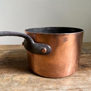 May include: A copper saucepan with a dark, riveted handle. The pot has a cylindrical shape and a dark interior. The handle is curved and attached to the side of the pot with a rivet. The pot is sitting on a wooden surface.