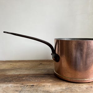 May include: A copper pot with a black handle and brass accents. The pot has a hammered finish and is sitting on a wooden surface.
