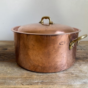 May include: A copper pot with a lid and brass handles. The pot is round with a slightly curved body and a matching lid. The handles are curved and attached to the sides of the pot. The pot is sitting on a wooden surface.
