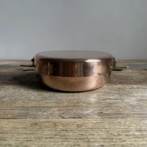 May include: A round copper pan with a flat lid and two small metal side handles. The pan has a polished, reflective surface and a smooth, cylindrical body. The design is simple and functional, showcasing the warm, metallic tone of the copper material.