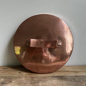 May include: A round copper pot lid with a rectangular handle riveted to the top. The lid has a polished, reflective surface and is set against a light-colored wall and a wooden surface.
