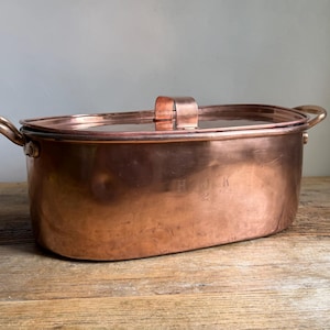 May include: An oval copper roasting pan with a matching lid, featuring two side handles and a central handle on the lid. The exterior surface of the copper pan displays a faint engraved inscription that reads H J K 2 on the front side of the vessel.