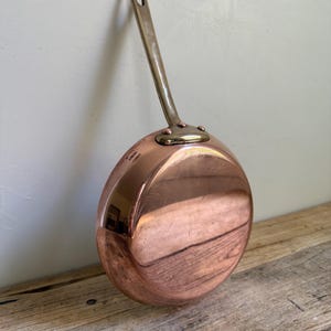 May include: A round copper frying pan with a polished metallic finish and a long, slender brass handle. The handle is securely attached to the side of the pan with three visible copper rivets. The surface shows light reflections and minor scratches.