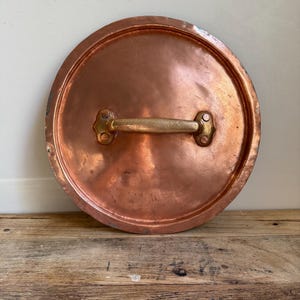 May include: A round copper pot lid with a brass handle. The lid has a polished finish and a slightly aged appearance. The handle is attached with rivets. The lid is likely for a cooking pot.