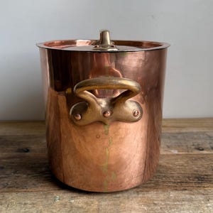 May include: A copper ice bucket with a lid and two handles. The bucket has a shiny, reflective surface and a brass handle on the lid. The handles are also brass. The bucket is cylindrical and sits on a wooden surface.