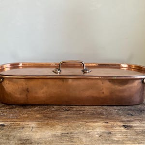 May include: Antique copper roasting pan with a lid and two handles. The pan is oval shaped and has a shiny finish.