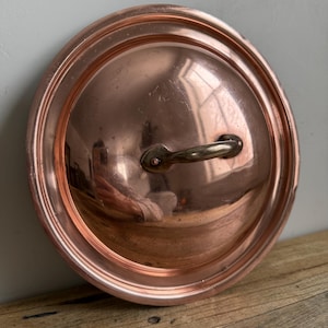 May include: A copper pot lid with a brass handle. The lid is round with a slightly raised rim and a polished finish. The handle is curved and attached to the lid with rivets. The lid is sitting on a wooden surface.