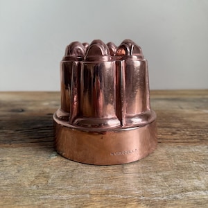 May include: A copper-colored, antique jelly mold with a decorative, fluted design. The mold has a rounded base and a series of rounded, dome-like shapes at the top. The text "HARRODS.L2" is stamped on the base. The mold is sitting on a wooden surface.