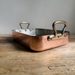 May include: A rectangular copper roasting pan featuring a metallic finish, a silver-toned interior lining, and two sturdy brass handles attached to the sides with visible rivets. The pan has rounded corners and a polished exterior surface.