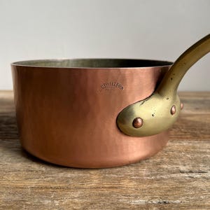 May include: A copper saucepan with a brass handle. The saucepan has a hammered finish and is stamped with the words "E. Dehillerin Paris".
