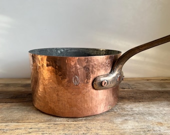May include: A hammered copper saucepan with a wooden handle. The saucepan has a distressed look and is a vintage kitchen item.