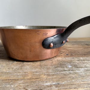 May include: Copper saucepan with a dark metal handle and rivets. The pan has a shiny copper exterior and a silver interior. The handle is attached with copper rivets. The words "MADE IN FRANCE" are stamped on the side of the pan.