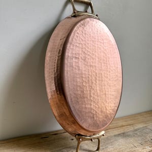 May include: Oval copper pan with hammered texture and brass handles. The pan is a warm copper color and has two brass handles on the sides. The pan is sitting on a wooden surface.