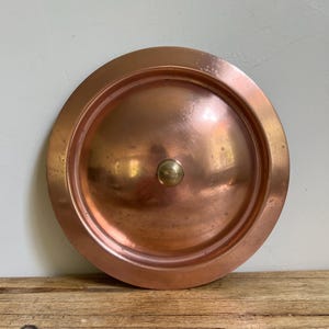 May include: A copper-colored, round lid with a central knob. The lid has a slightly domed center and a raised rim. The metal has a warm, reddish-brown hue and shows signs of age. The knob is a lighter, brassy color.