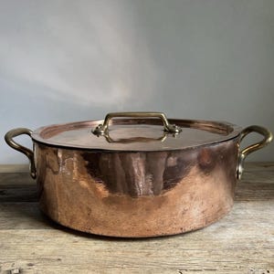 May include: A copper cooking pot with a lid and brass handles. The pot is round with a hammered texture, reflecting light. The lid has a brass handle. The pot sits on a wooden surface.