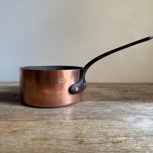 May include: A round copper saucepan with a smooth, polished exterior and a long, dark, curved iron handle attached with two copper rivets. The side of the pan features a small, oval-shaped metal stamp with legible text, showcasing a classic design.