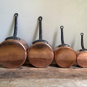 May include: Four copper saucepans of varying sizes with black handles. The pans have a shiny, reflective surface and are arranged in a row on a wooden surface. The handles are curved and have a loop at the end.