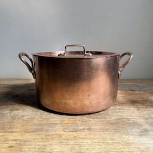 May include: A copper cooking pot with a lid, featuring two side handles and a top handle. The pot has a shiny, reflective surface and sits on a wooden surface. The pot is a warm copper color.