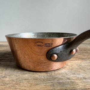 May include: A copper saucepan with a dark metal handle. The pan has a shiny copper exterior and a silver interior. The handle is riveted to the pan. The text "MADE IN FRANCE" is visible on the side of the pan.