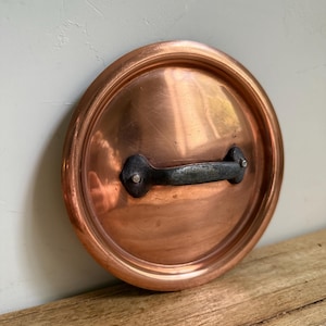 May include: A copper pot lid with a dark metal handle. The lid is round and has a slightly raised rim. The handle is riveted to the lid. The lid is sitting on a wooden surface.
