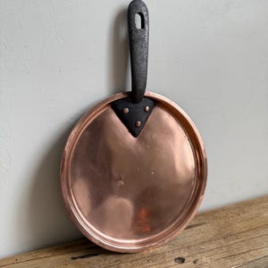 May include: A copper-colored, round pan with a dark metal handle. The handle has a hole at the top for hanging. The pan is sitting on a wooden surface.