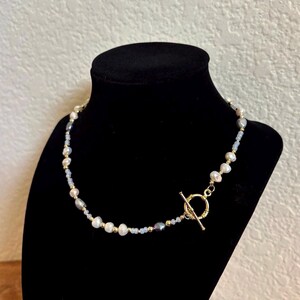 May include: A delicate necklace featuring a strand of white and gray pearls interspersed with small, light blue beads. The necklace has gold-colored accents and a gold toggle clasp. The jewelry is displayed on a black velvet bust.