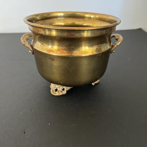 May include: A polished brass bowl featuring a wide, rounded rim, two small decorative side handles, and three ornate, scrolled feet. The metal surface displays a warm, metallic gold tone with subtle variations in its reflective finish.