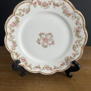 May include: A white porcelain plate with a scalloped edge and a gold-trimmed rim. The plate features a delicate floral pattern of pink roses and green leaves arranged in a circular border around the edge and a small cluster in the center.