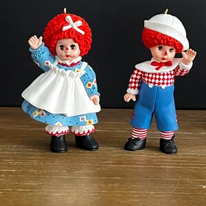May include: Two decorative figurines featuring a doll with bright red hair, a blue floral dress, and a white apron, alongside a doll with red hair, a white sailor hat, a red and white checkered shirt, and blue shorts, both with black shoes.