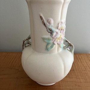 May include: A cream-colored ceramic vase with a floral design. The vase features a bulbous base and a flared top with a scalloped edge. Delicate pink and yellow flowers with green leaves and brown stems adorn the front. The vase has two decorative handles.