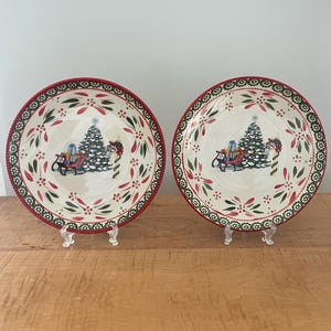 May include: Two round Christmas plates with a cream-colored center featuring a decorated Christmas tree, gifts, and a sleigh. The plates have a red and green border with a decorative pattern. The plates are displayed on clear stands.