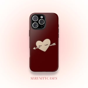 May include: A burgundy phone case with a heart pierced by an arrow. The heart is light tan with closed eyes. The case has a glossy finish and a camera module with three lenses. The text "SERENITYCASES" is at the bottom of the case.