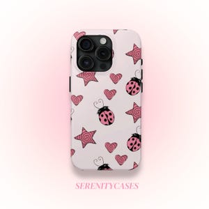 May include: White phone case with a repeating pattern of pink stars, hearts, and ladybugs. The case has a black camera module and the text "SERENITYCASES" at the bottom.