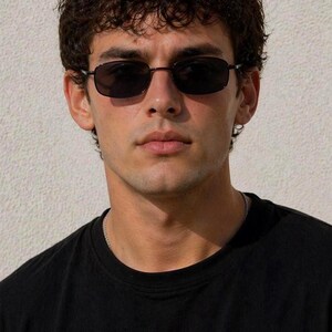 May include: Black rectangular sunglasses with dark lenses. The sunglasses have a thin, dark frame. The person in the image is wearing a black t-shirt and a silver chain necklace.