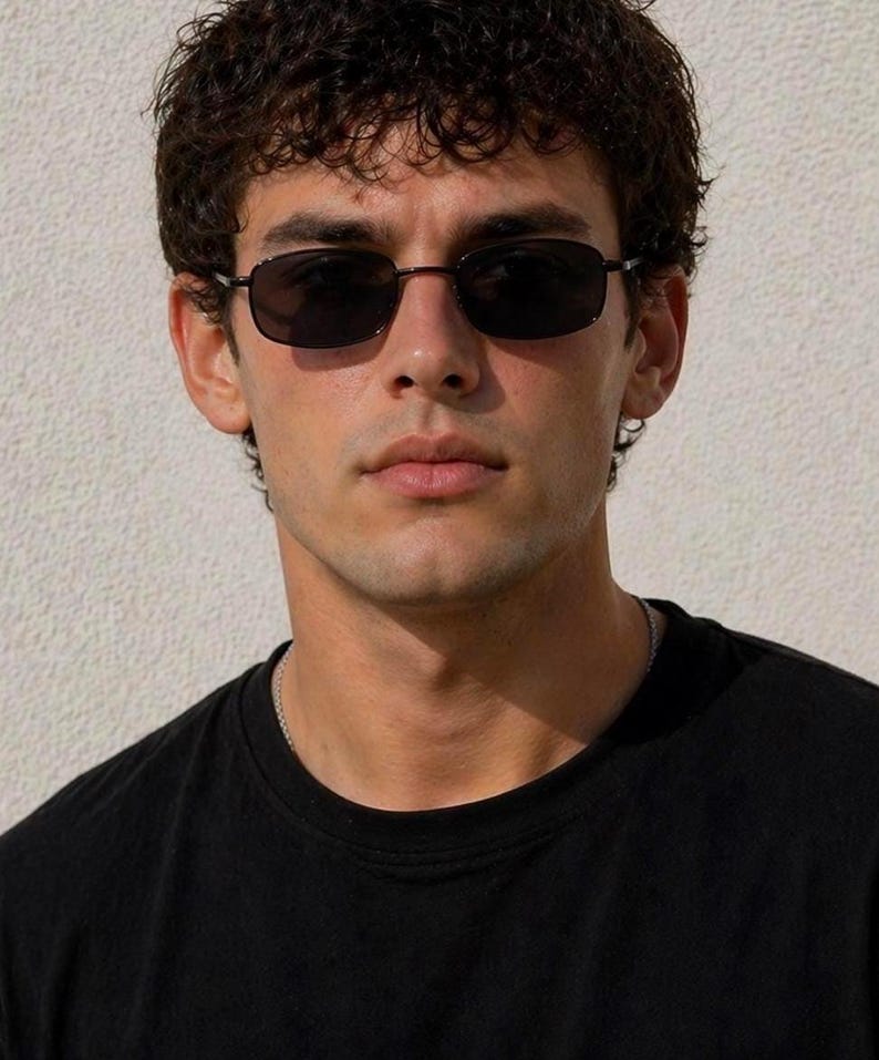 May include: Black rectangular sunglasses with dark lenses. The sunglasses have a thin, dark frame. The person in the image is wearing a black t-shirt and a silver chain necklace.