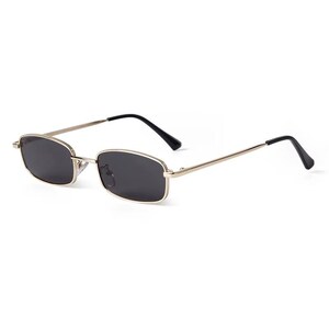 May include: Rectangular sunglasses with a gold-colored frame and dark lenses. The temples are thin and silver, with black tips. These sunglasses offer a classic, minimalist design.
