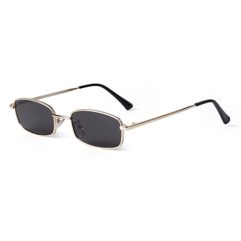May include: Rectangular sunglasses with a gold-colored frame and dark lenses. The temples are thin and silver, with black tips. These sunglasses offer a classic, minimalist design.