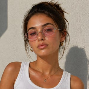 May include: A woman wearing silver-framed eyeglasses with pink lenses, gold hoop earrings, and a gold pendant necklace. She is wearing a white tank top. The background is a plain white wall.