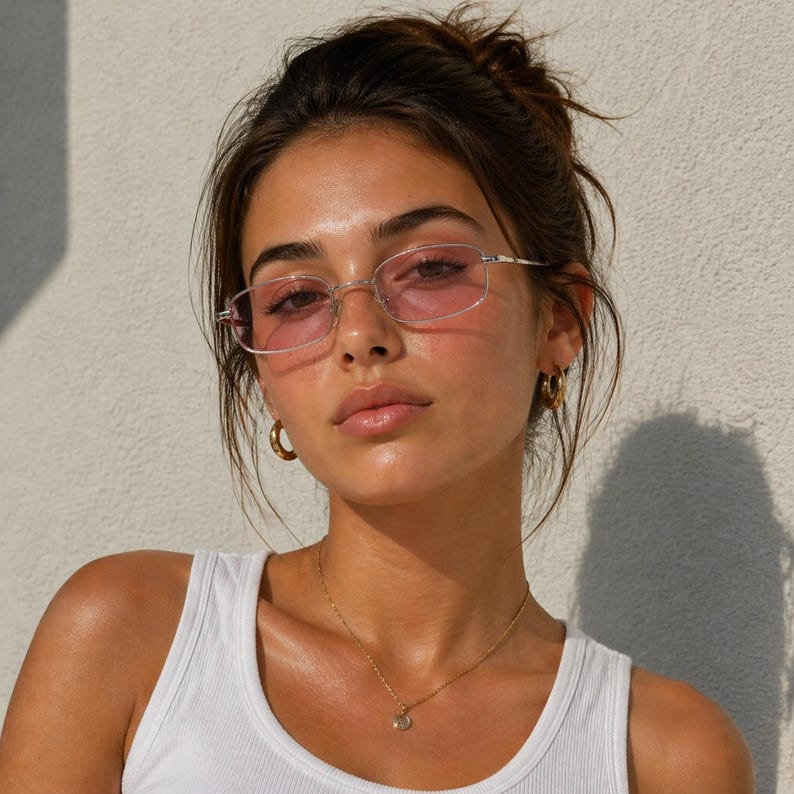May include: A woman wearing silver-framed eyeglasses with pink lenses, gold hoop earrings, and a gold pendant necklace. She is wearing a white tank top. The background is a plain white wall.
