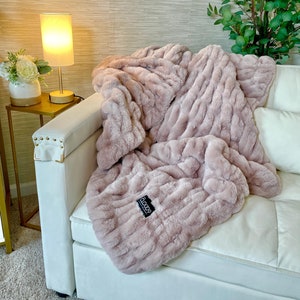 May include: A soft, plush, pink faux fur throw blanket draped over a white couch. The blanket has a ribbed texture and a black label with the word "Cozy" on it.