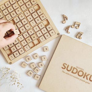 Könnte beinhalten: Holz-Sudoku-Spiel mit Zahlensteinen und einem Holzbrett. Das Spiel ist auf einer hellbraunen Oberfläche mit ausgeschnittenen Musiknoten angeordnet.