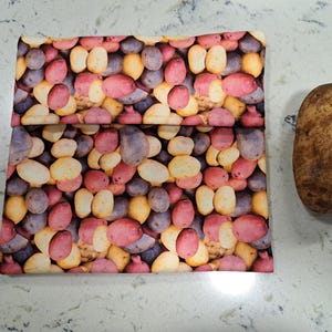 May include: A reusable food storage bag with a pattern of colorful potatoes. The bag is folded in half and is lying on a white countertop. A single brown potato is sitting to the right of the bag.