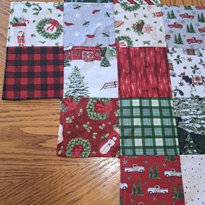 May include: A patchwork Christmas table runner featuring diverse festive patterns. The design includes red and black buffalo check, red trucks, snow-covered barns, wreaths, and Christmas trees. The runner is made of fabric and is placed on a wooden surface.