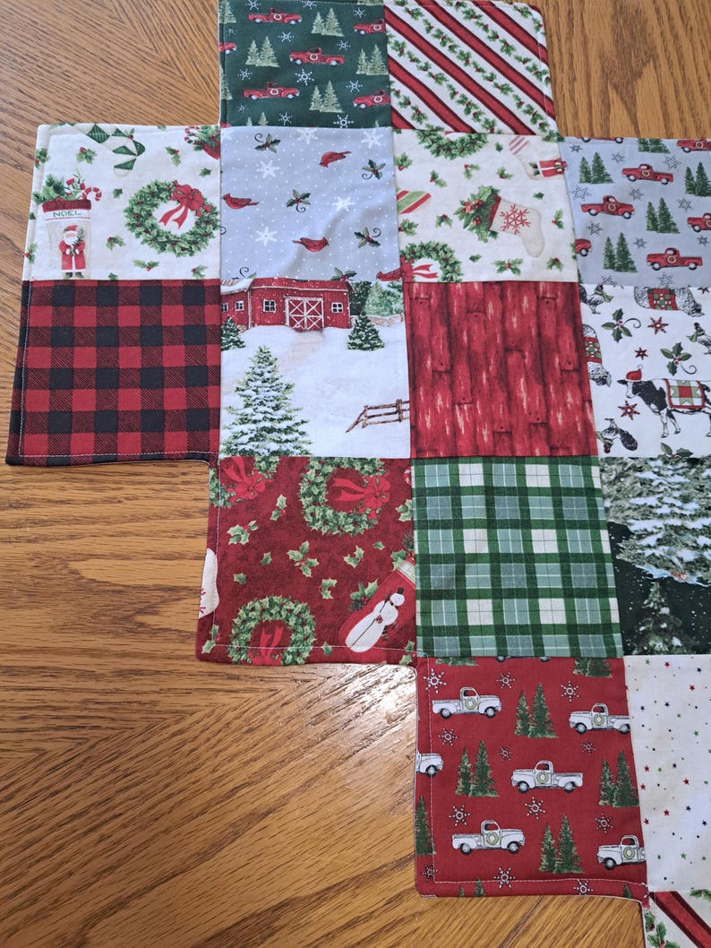 May include: A patchwork Christmas table runner featuring diverse festive patterns. The design includes red and black buffalo check, red trucks, snow-covered barns, wreaths, and Christmas trees. The runner is made of fabric and is placed on a wooden surface.