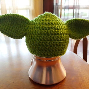 May include: Green crocheted hat with large ears, resembling a character from the Star Wars franchise. The hat is sitting on a silver stand.