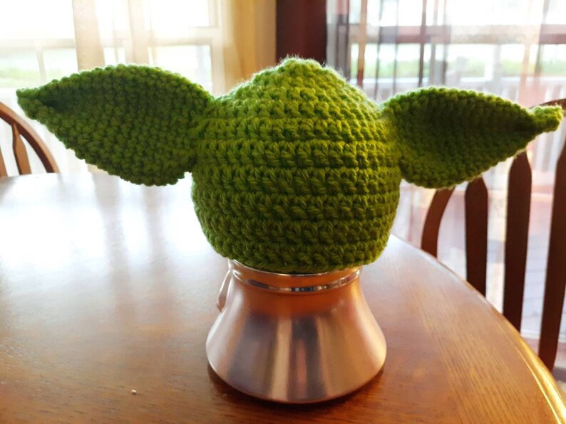 May include: Green crocheted hat with large ears, resembling a character from the Star Wars franchise. The hat is sitting on a silver stand.