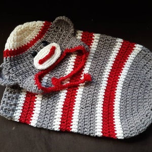 May include: A handmade crochet sock monkey baby blanket. The blanket features alternating stripes of red, white, and gray. The monkey's face and hat are visible, with black button eyes and a red mouth.