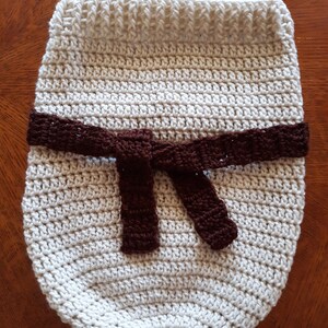 May include: A crocheted baby blanket in a cream color with a brown crocheted belt tied in a bow.